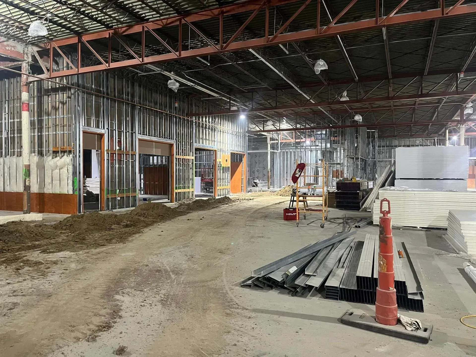 An indoor construction site showing metal wall framing, dirt floors, stacked building materials, and an orange safety cone.