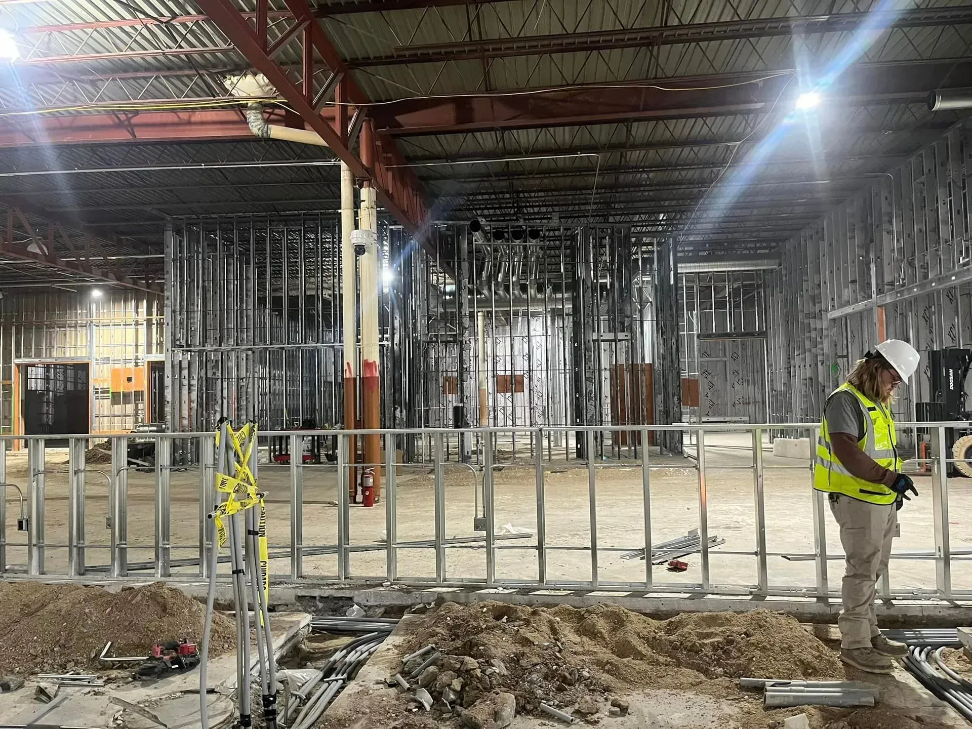 A person wearing a hard hat and safety vest standing in an indoor construction site with metal wall framing and debris.