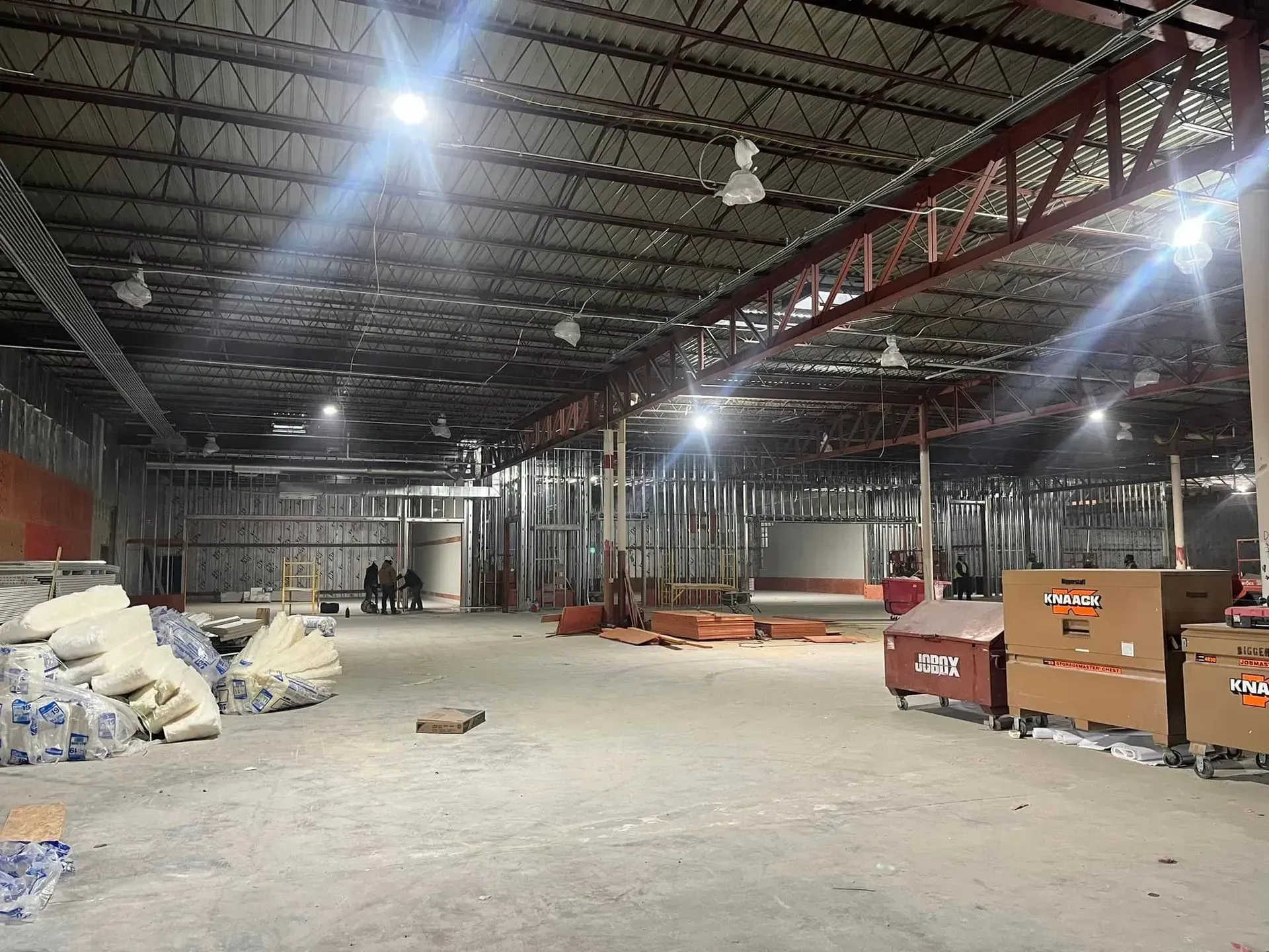 An interior construction site features metal framing, an open ceiling with overhead lighting, and stacked boxes.