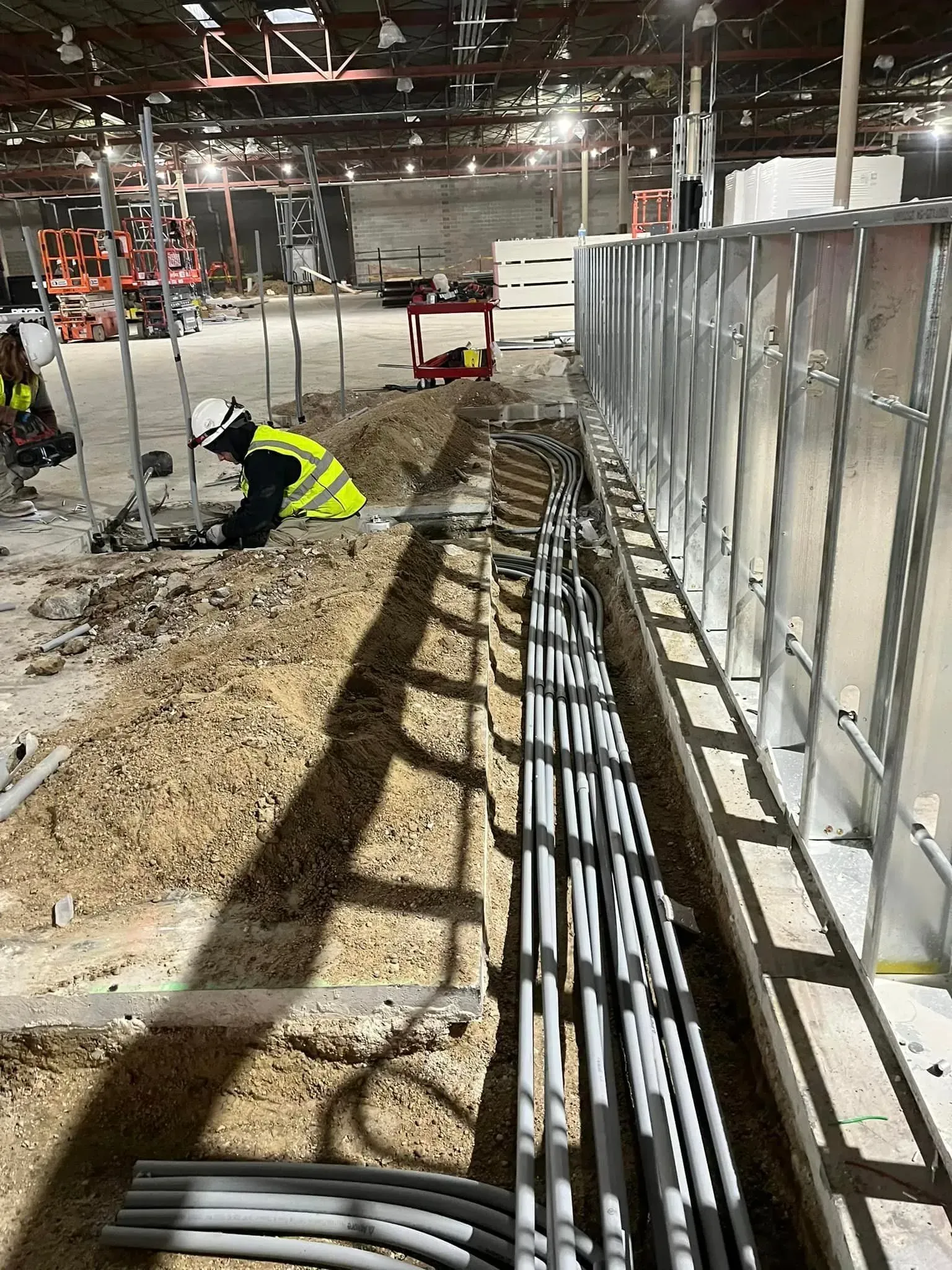 A construction worker in a high-visibility vest kneels on a dirt floor, installing electrical conduits near metal walls.