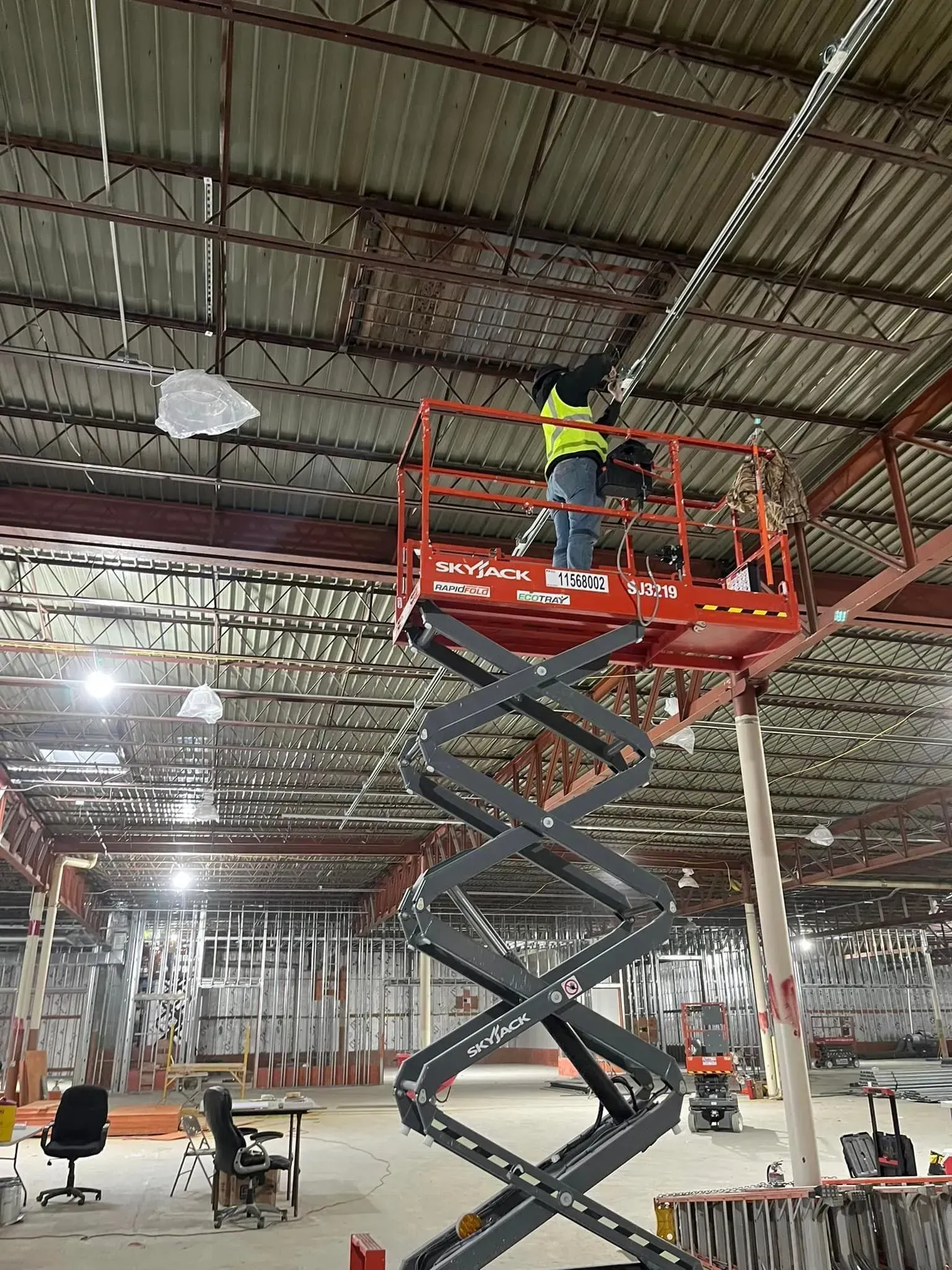 A person in a yellow safety vest works on the ceiling while standing on a tall, red scissor lift in an industrial space.