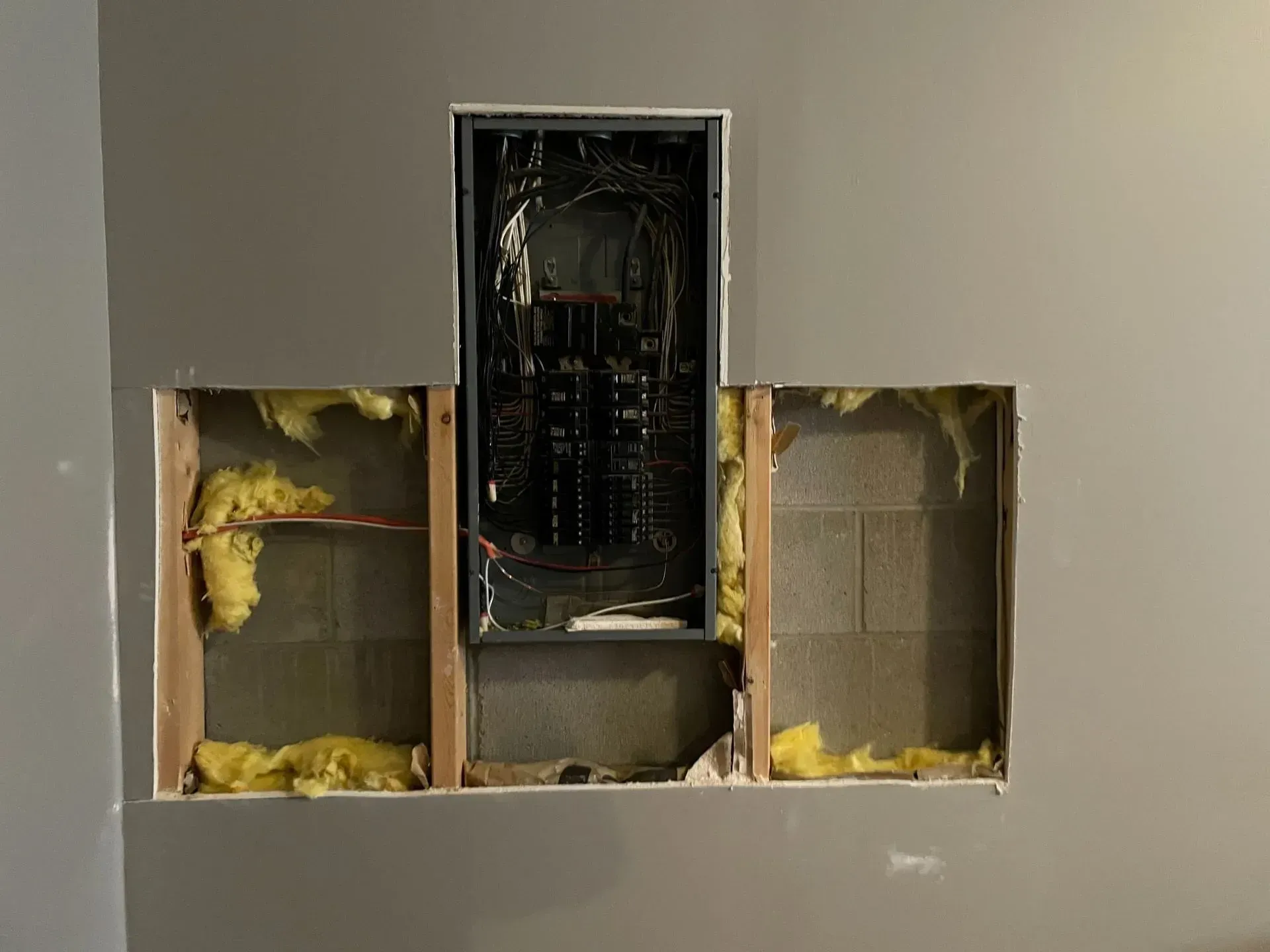 An electrical breaker panel centered in a rectangular hole in a gray wall, revealing wooden studs and yellow insulation.