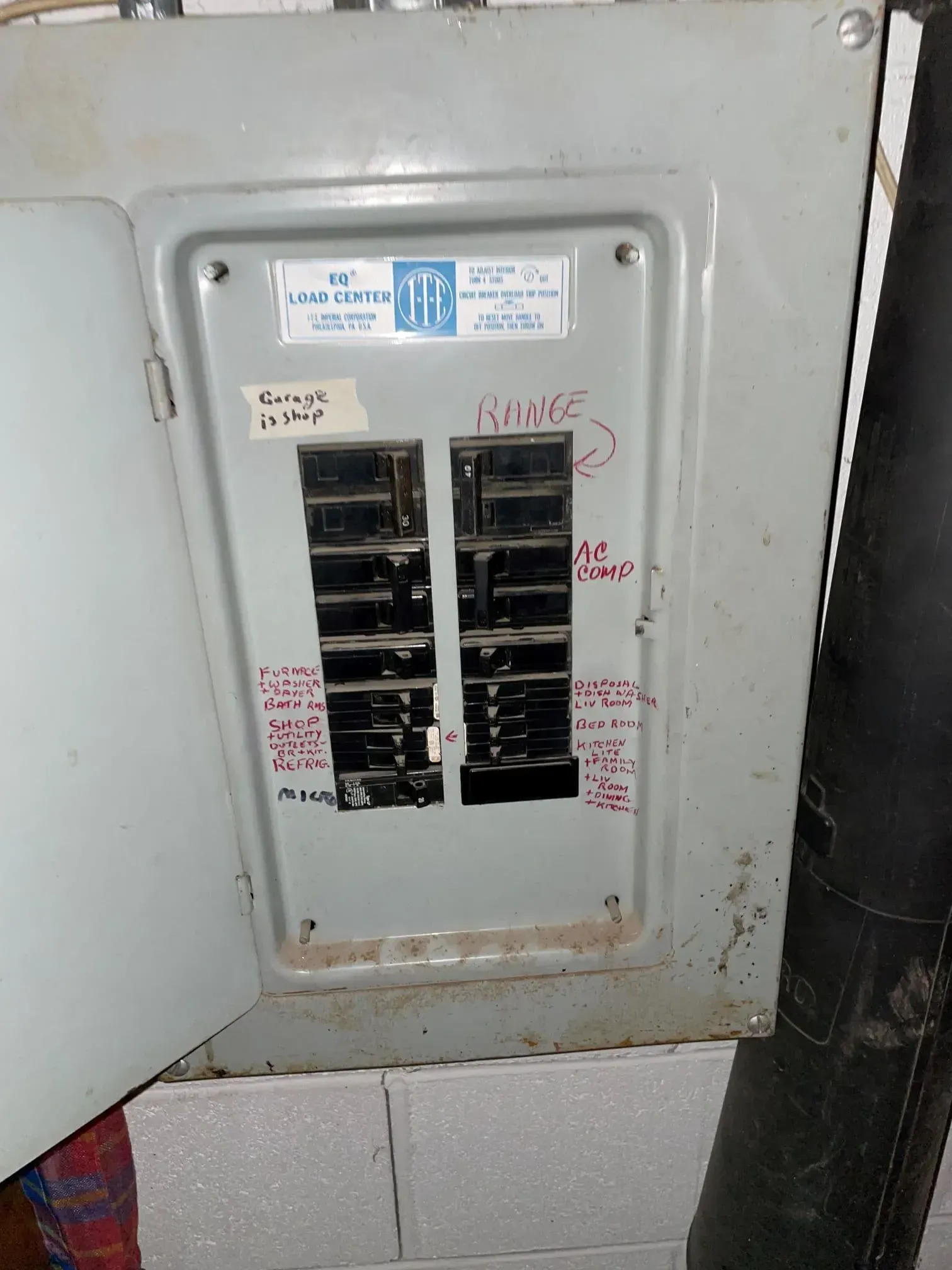 A grey, open electrical breaker panel mounted on a cinder block wall with handwritten labels on the door and panel.