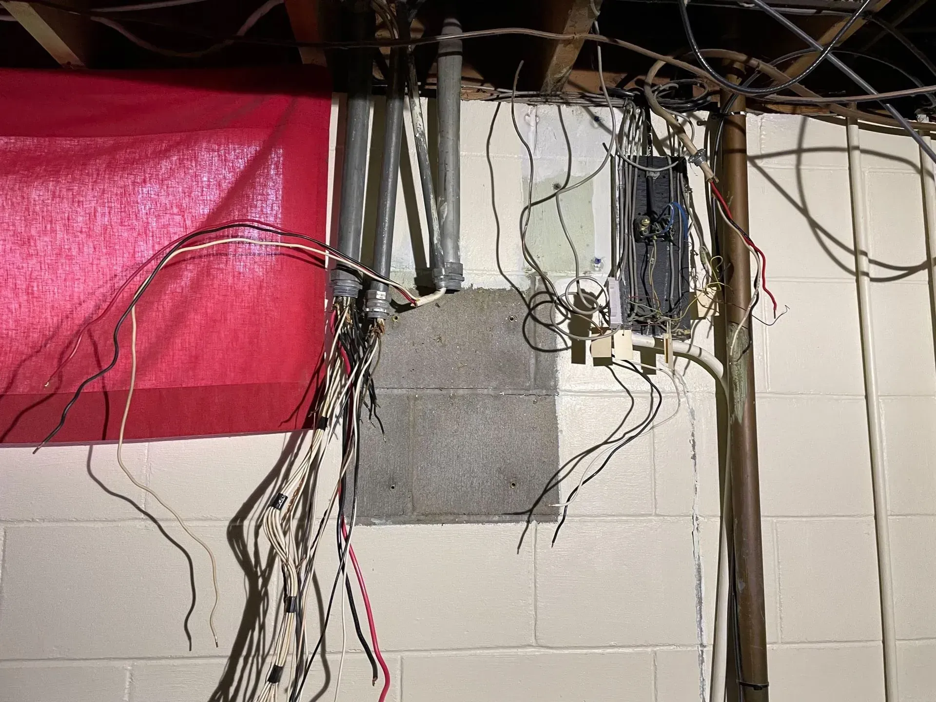 Exposed electrical wiring and conduit pipes run along a cinder block basement wall with a partially open electrical panel.