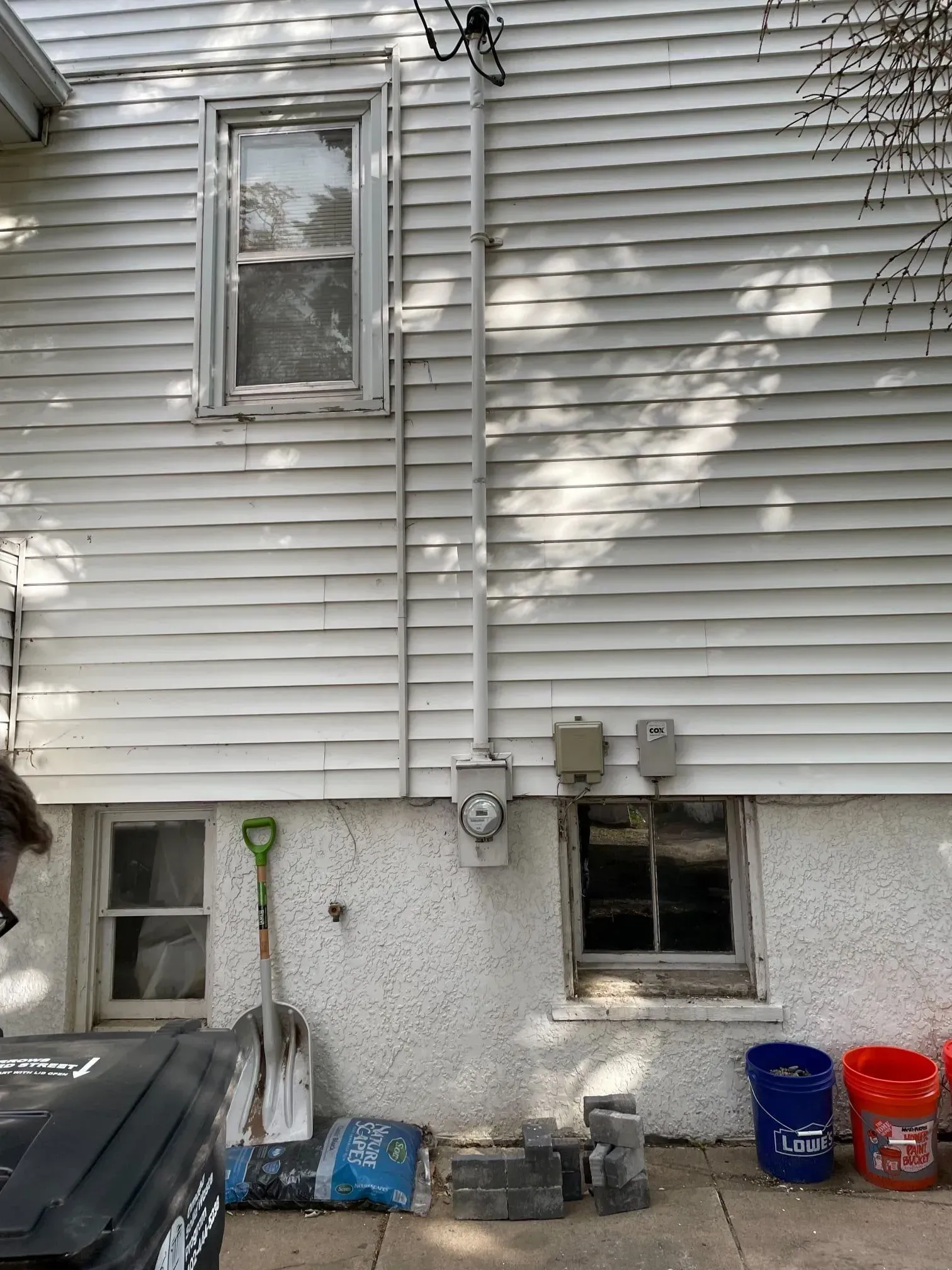 Exterior view of a white-sided house with a basement foundation, two lower windows, an electrical meter, and tools.