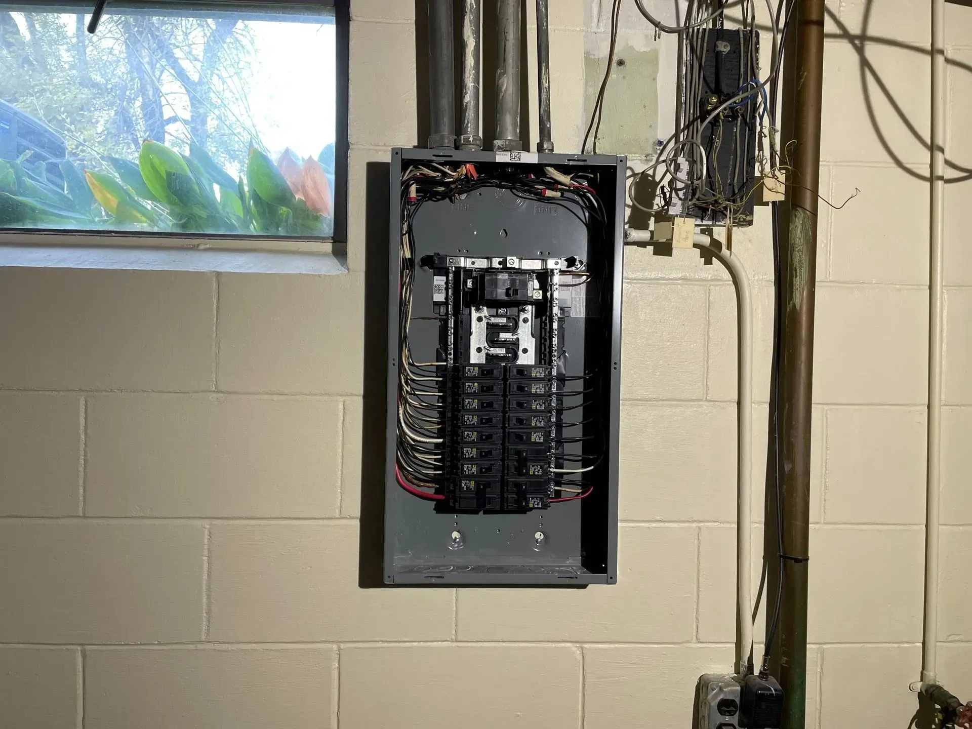 An open electrical panel box mounted on a concrete block wall next to a window, with exposed wiring and breakers inside.