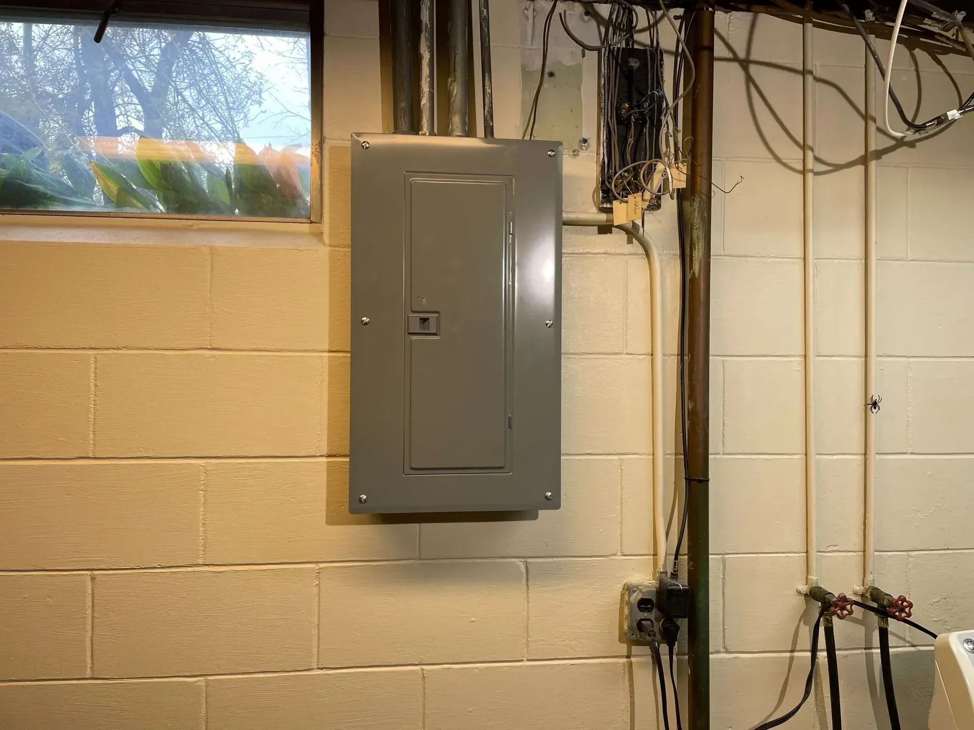 A grey electrical panel mounted on a beige cinder block wall in a basement next to a small window and exposed wiring.