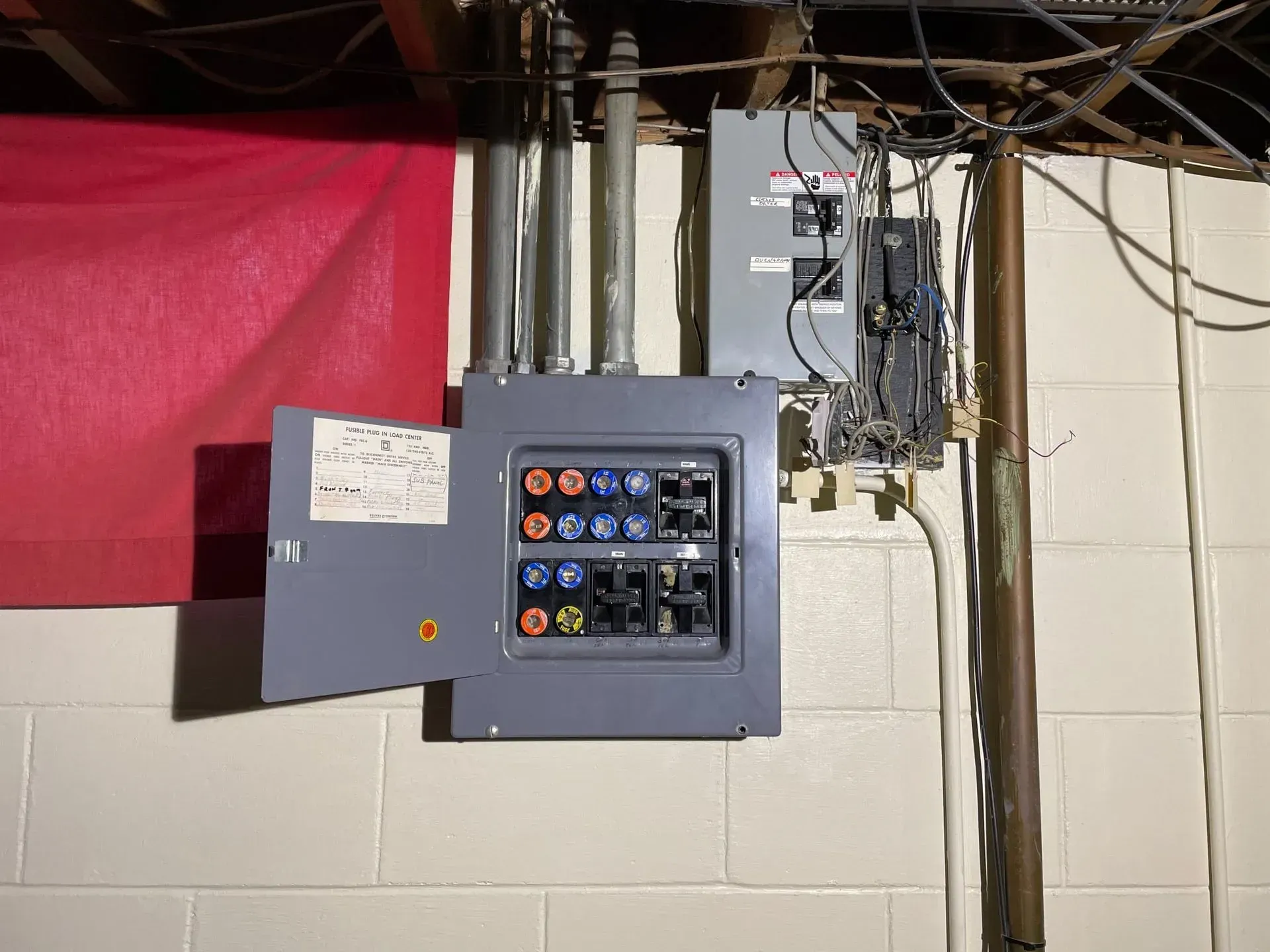 A gray electrical service panel with fuses and an attached subpanel mounted on a beige cinder block wall.