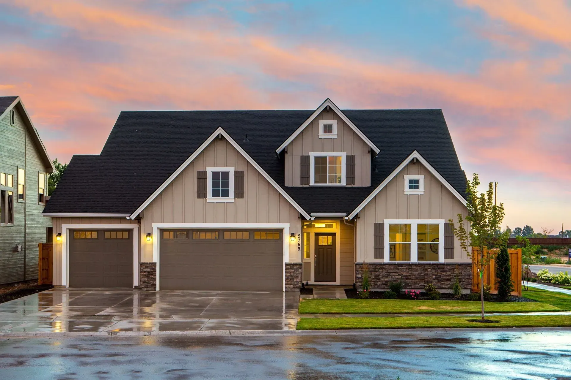 A tan, two-story suburban home with a three-car garage and stone accents under a colorful sunset sky.