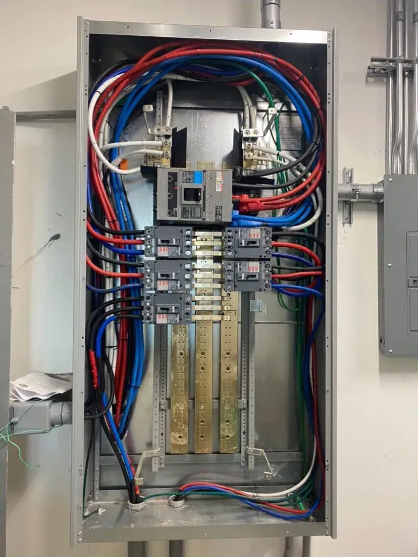 Open electrical distribution panel showing circuit breakers, copper busbars, and multi-colored wiring inside a metal box.