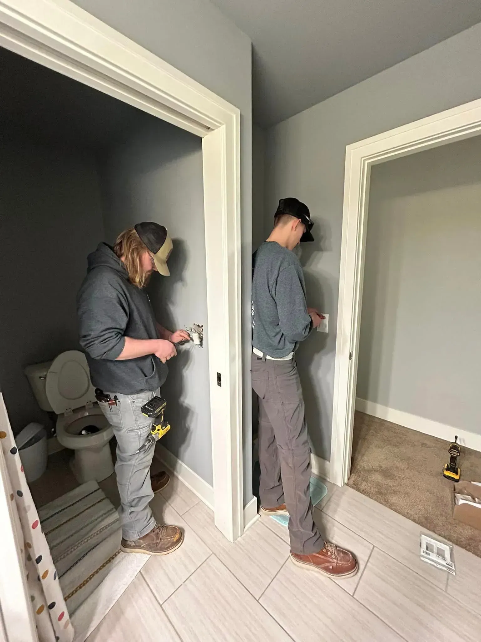 Two workers in grey hoodies and jeans perform electrical work on wall outlets in a house with grey walls.