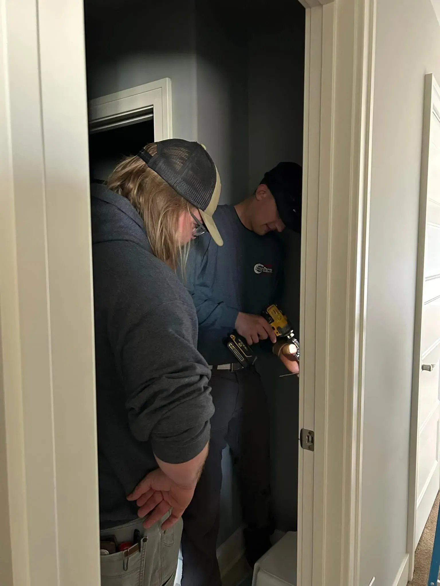 Two people work on a wall fixture in a narrow doorway, one holding a power drill while the other looks on.
