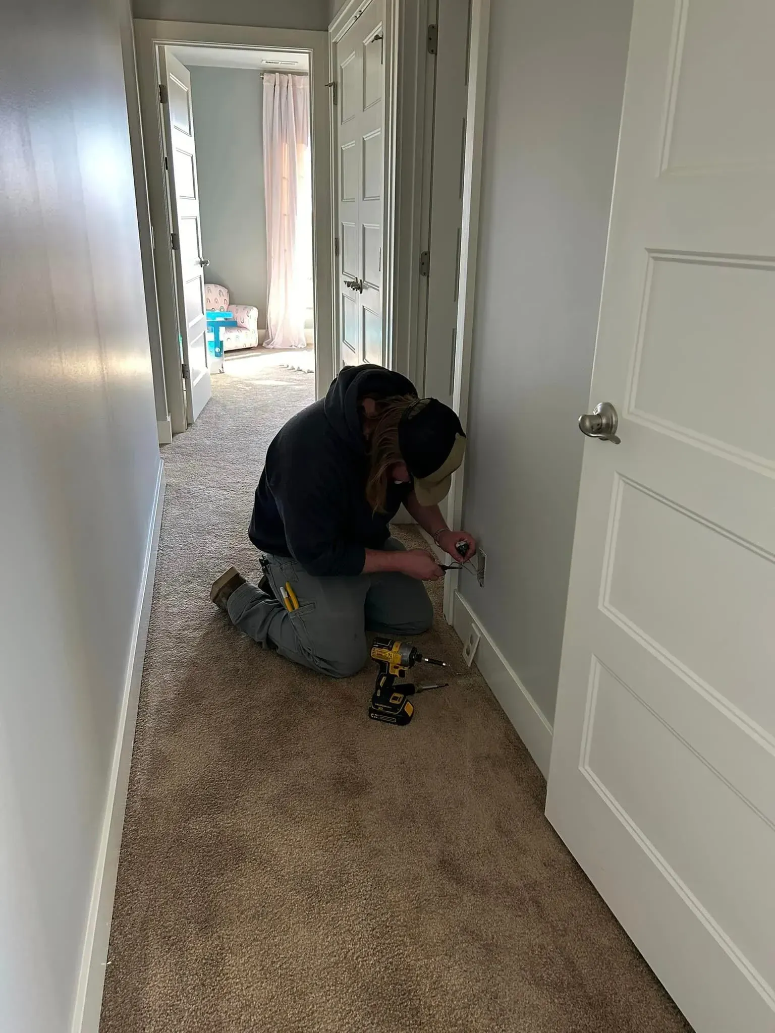 A person in work clothes kneels on a carpeted hallway floor, using a tool on a wall outlet near a white door.