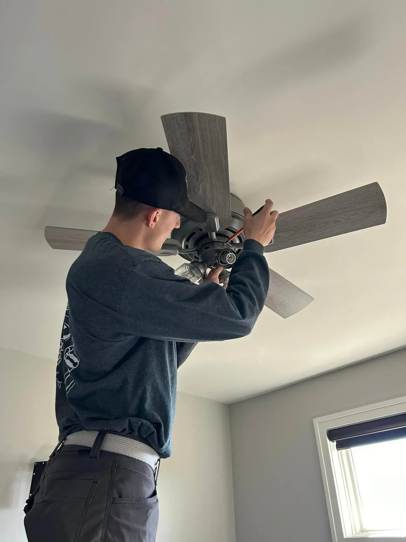 A person wearing a baseball cap and a dark long-sleeved shirt standing on a ladder while installing a ceiling fan.