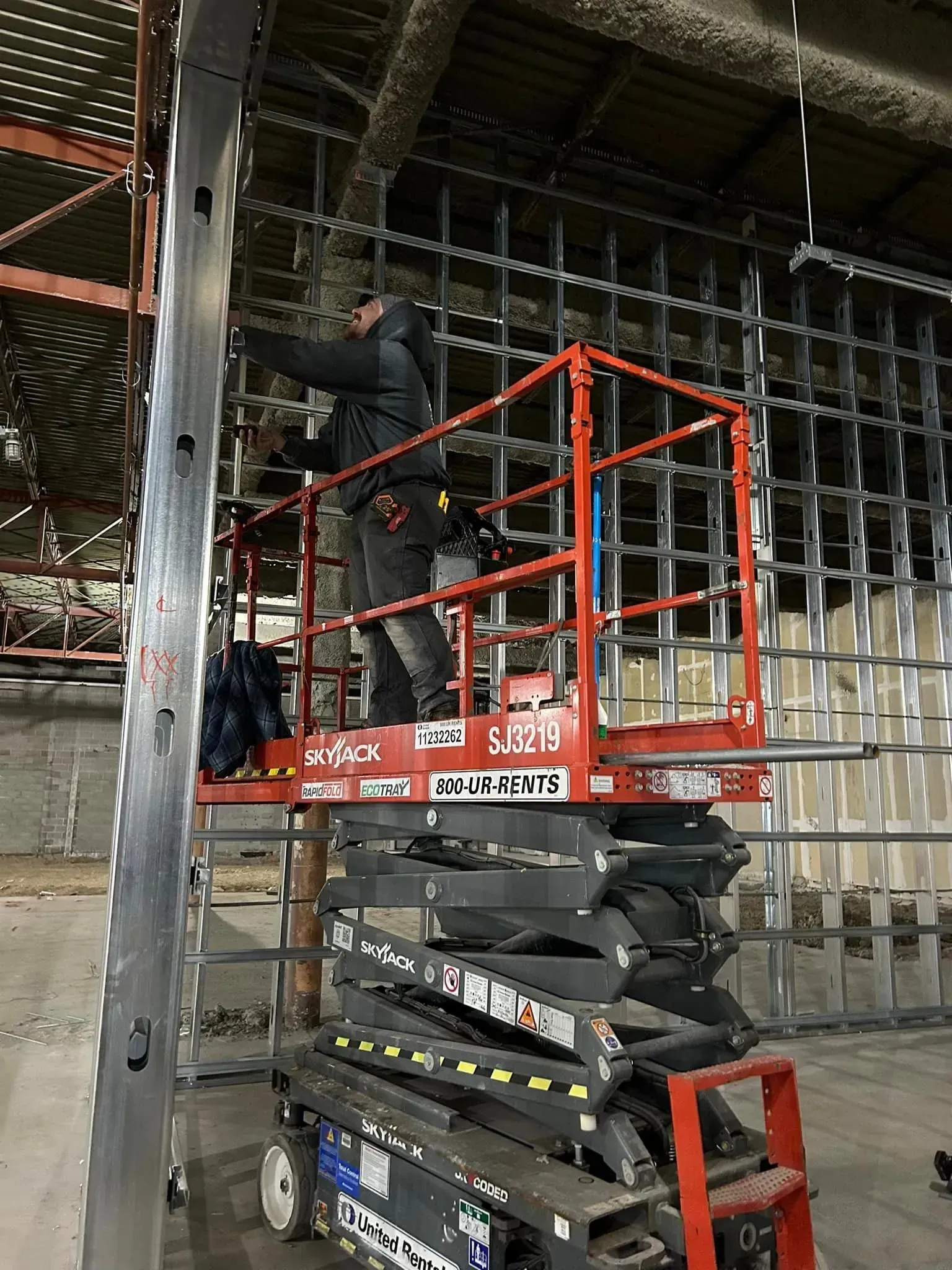A person in dark work clothes stands on a red scissor lift, working on a metal wall frame inside an unfinished building.