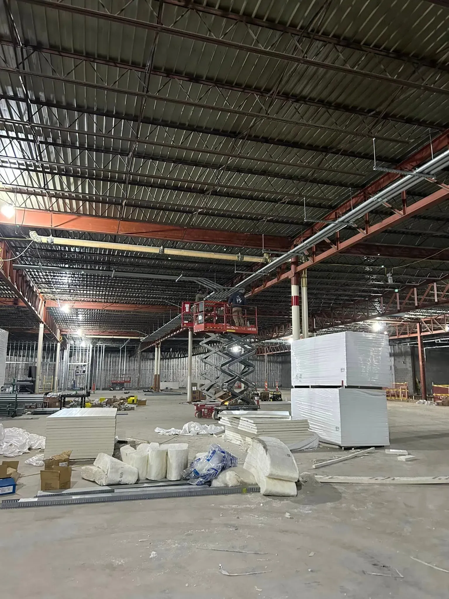 Construction interior with a high, metal-decked ceiling, exposed steel beams, stacks of white insulation, and debris.