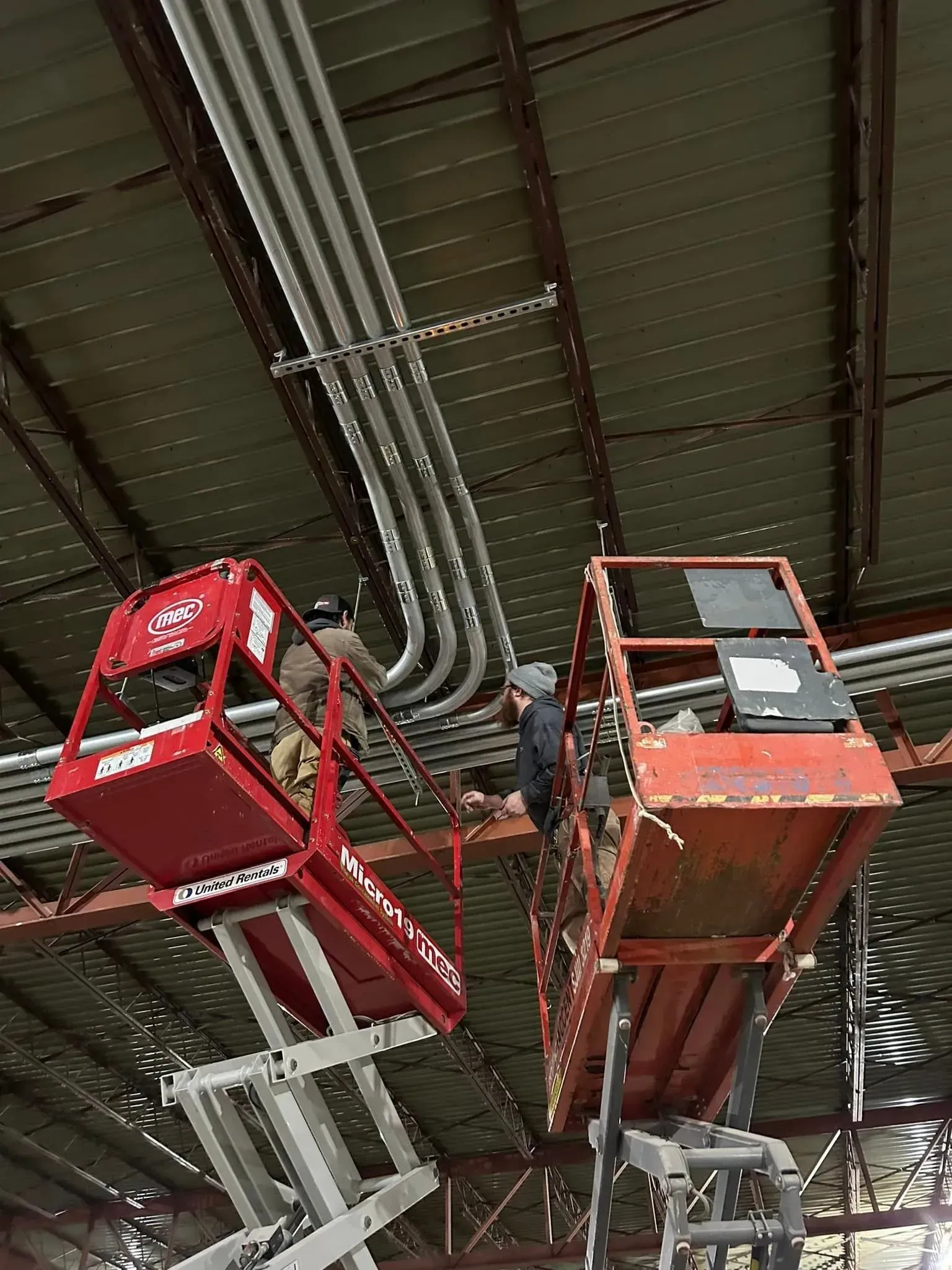 Two people on elevated red scissor lifts work on overhead electrical conduits in an industrial warehouse.