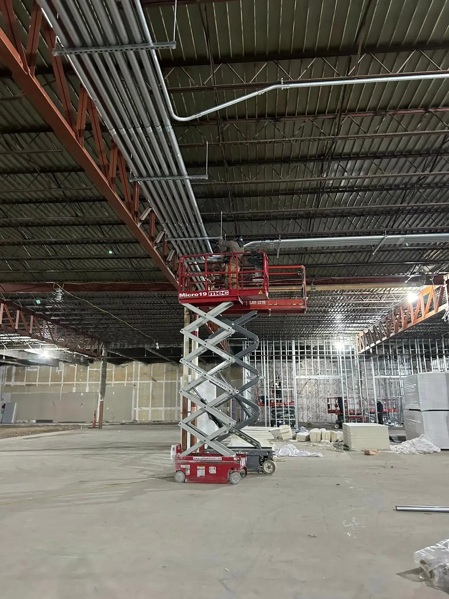 A red scissor lift with a worker on the platform is positioned under a ceiling with extensive conduit and metal trusses.