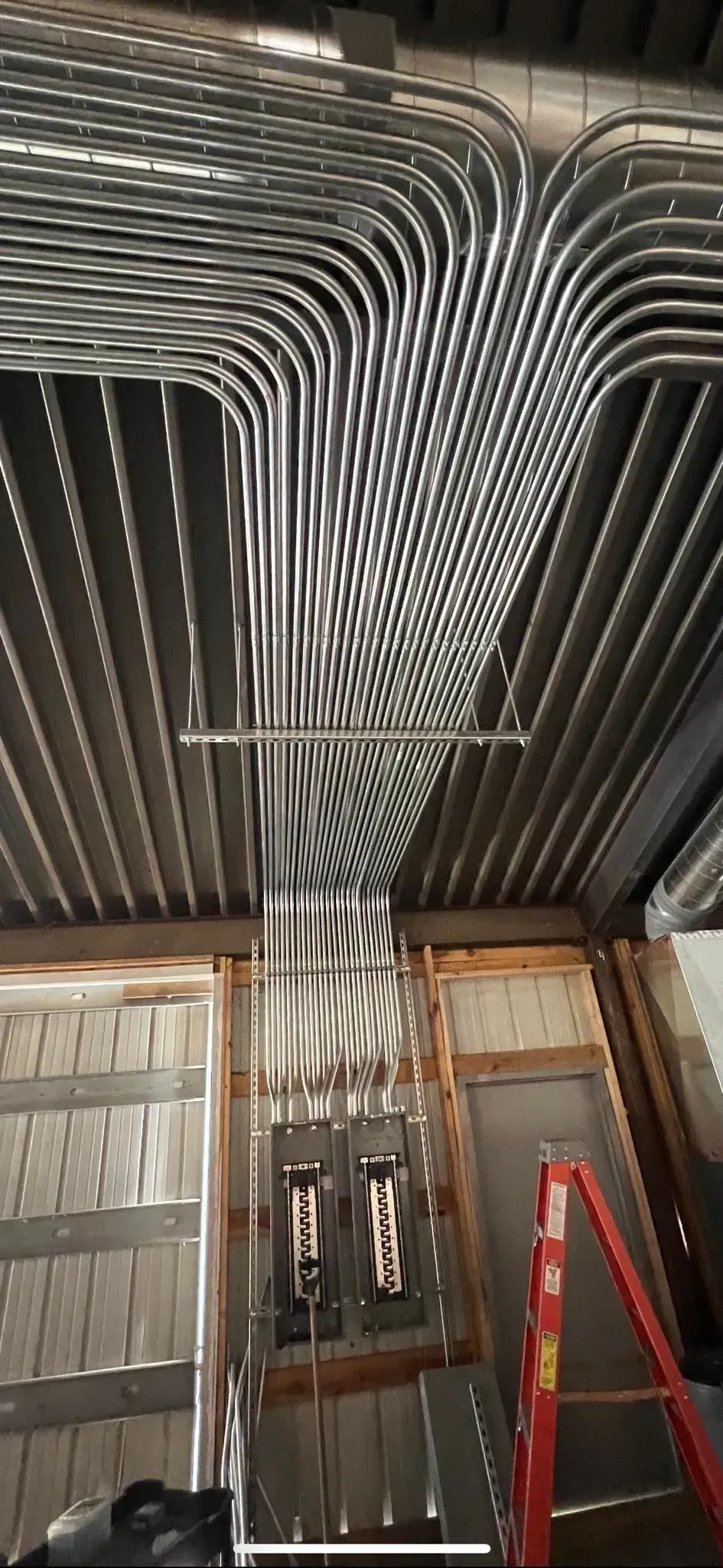 A dense array of silver electrical conduits spanning a ceiling and running down into a wall-mounted electrical panel.