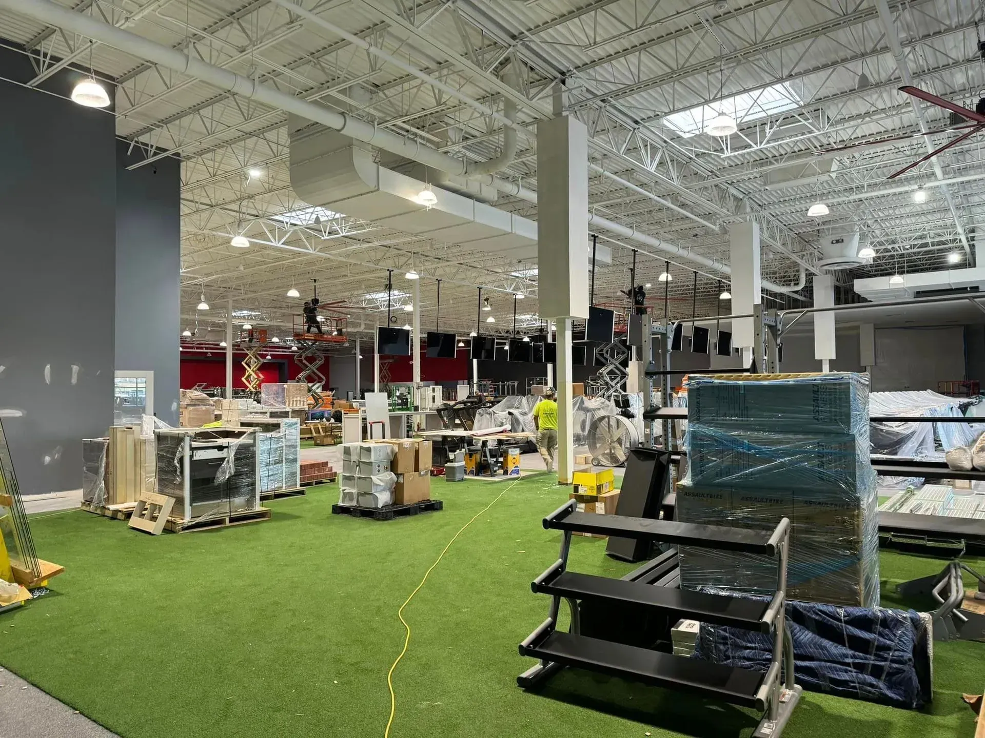 An indoor warehouse space under construction with green artificial turf flooring, equipment, and shelves.