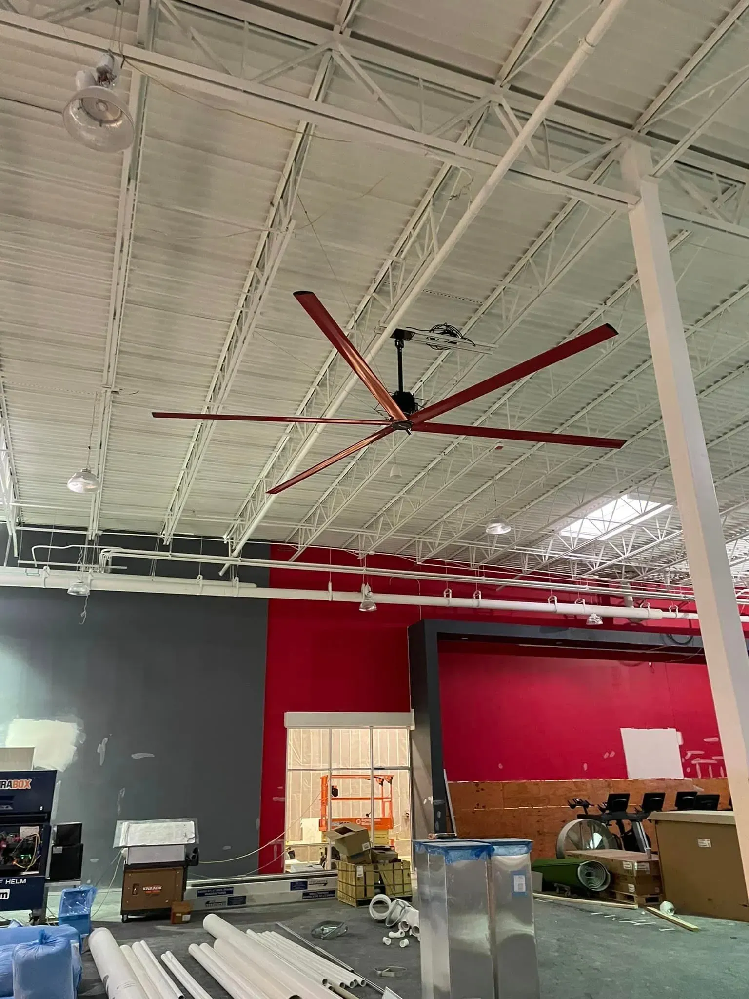 A large red industrial ceiling fan hangs in a spacious, unfinished room with a white metal ceiling and red-painted walls.