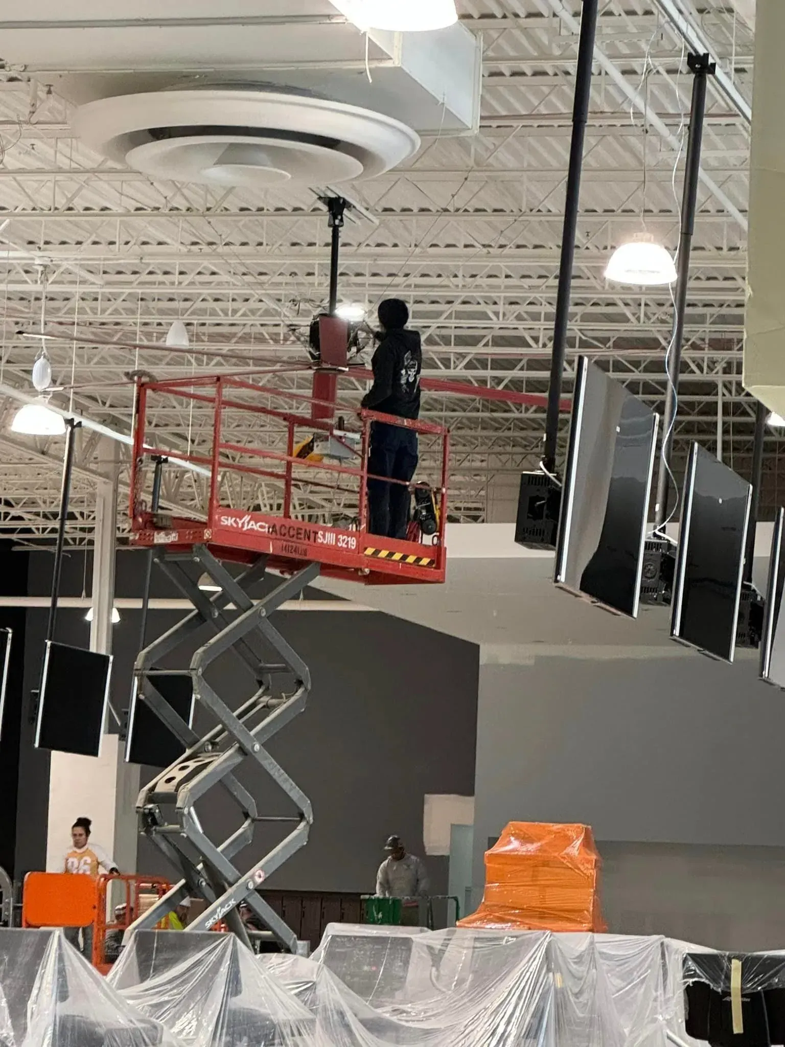 A person stands on a red scissor lift, installing large black panels suspended from an industrial ceiling.