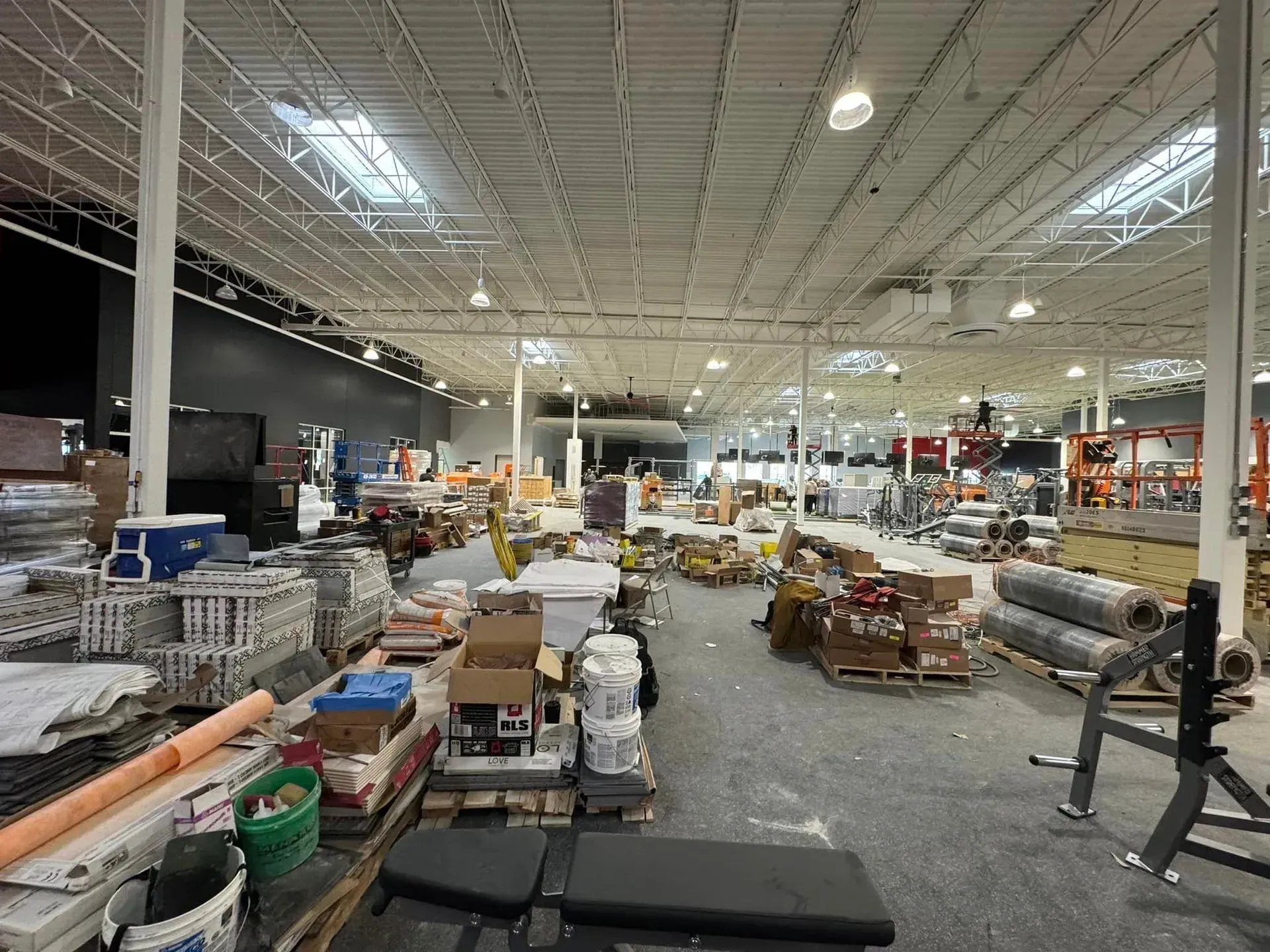 A cluttered interior of a large, high-ceilinged warehouse or retail space under construction, with pallets and supplies.