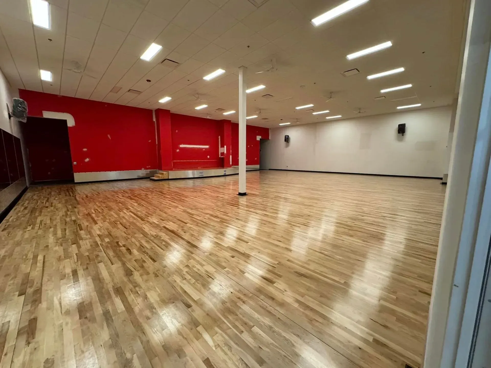 An empty indoor fitness studio with hardwood floors, a bright red accent wall, a support pillar, and overhead lighting.