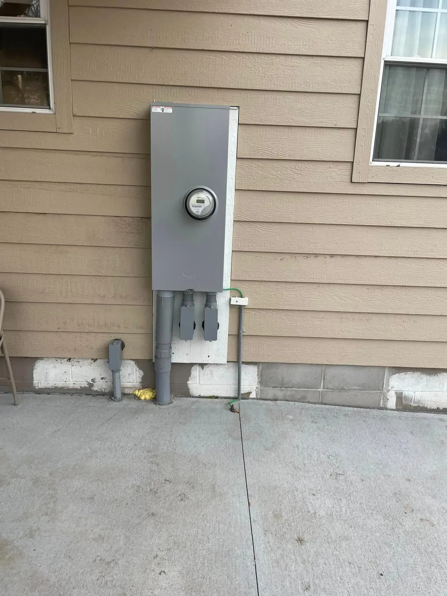 A gray electrical meter box mounted on the side of a tan house with a concrete slab foundation and a nearby window.