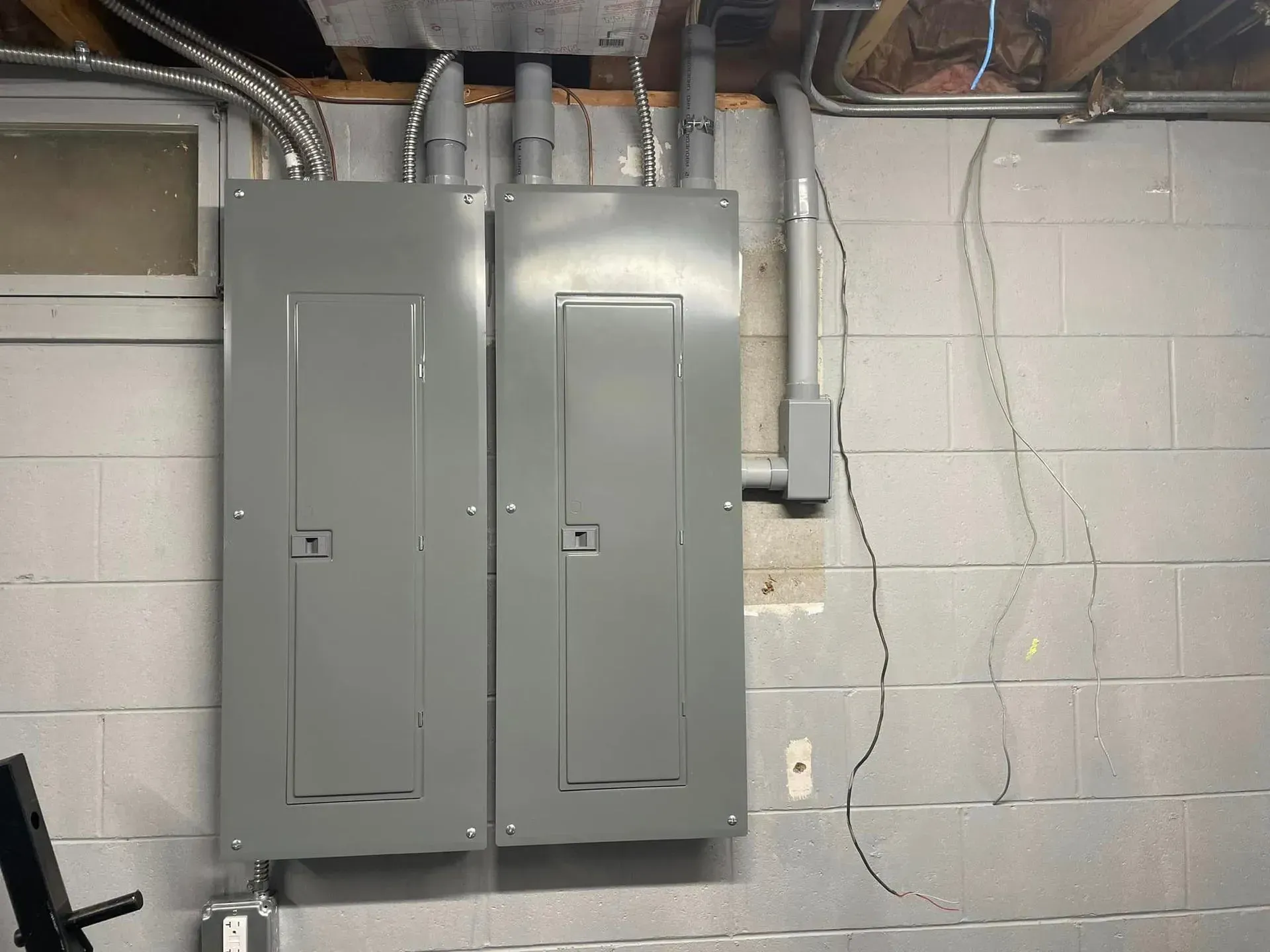 Two gray electrical breaker panels mounted side-by-side on a cinder block wall, with conduits and loose wires above.