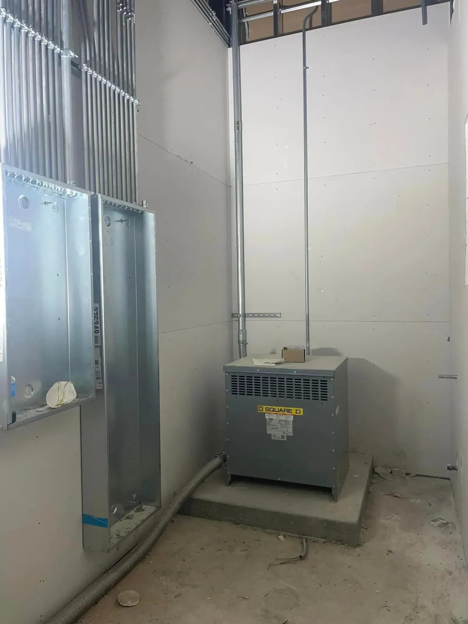 An electrical transformer sits on a concrete pad in a room with unfinished drywall and wall-mounted electrical conduits.
