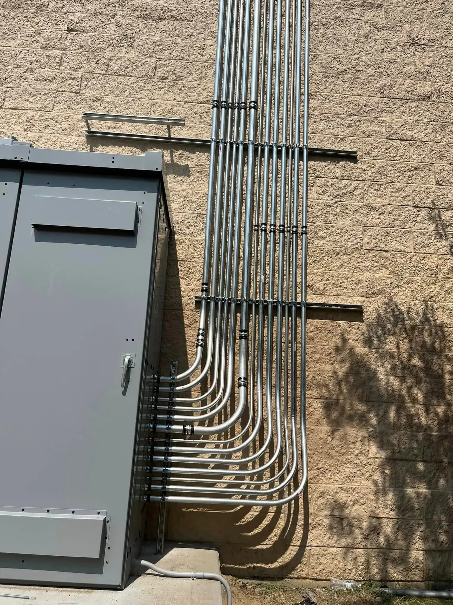 Multiple metal electrical conduits neatly routed along a textured exterior wall into a grey metal utility box.