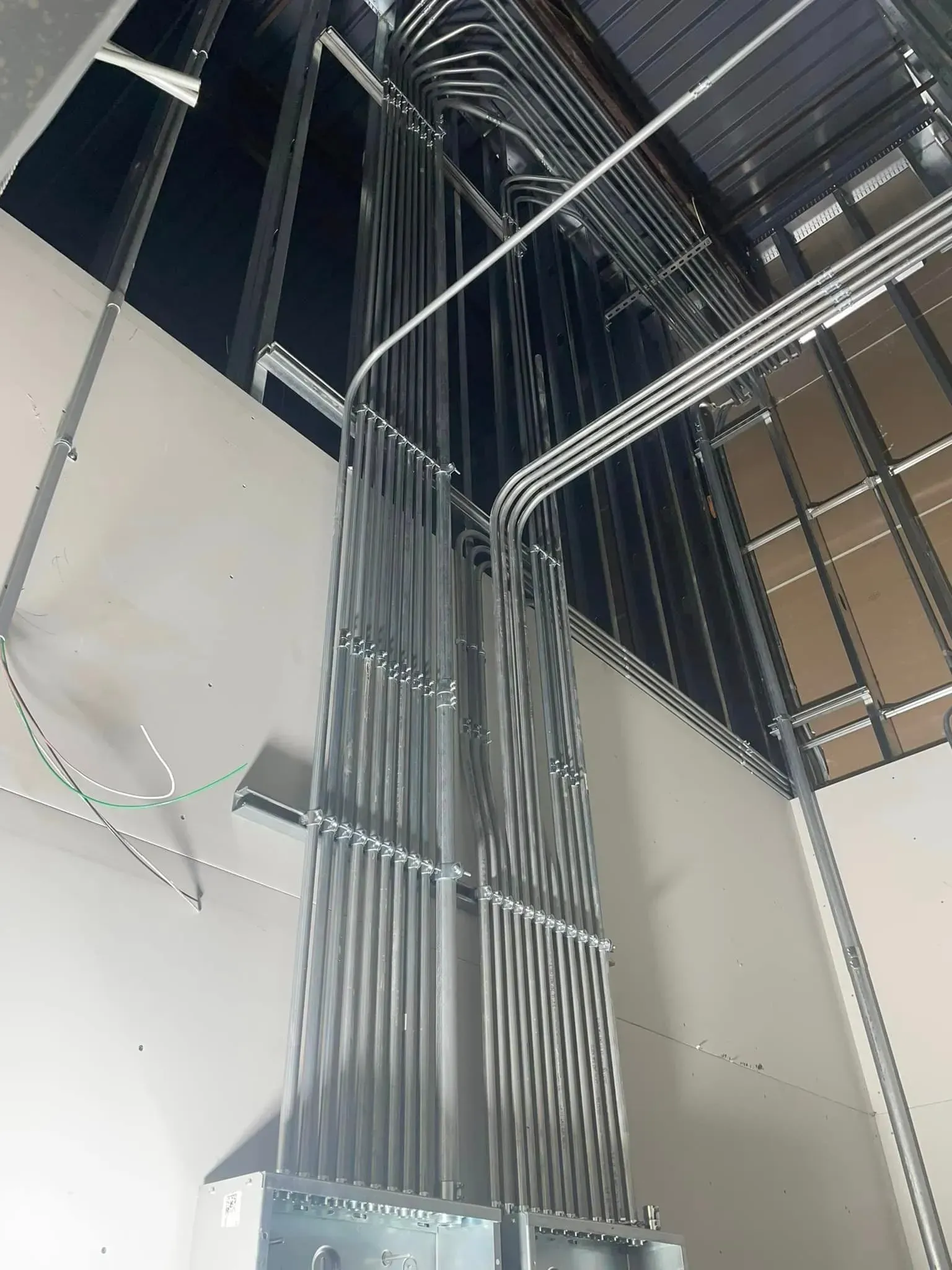 A vertical view of numerous metallic electrical conduits running up a wall from a junction box toward a ceiling frame.