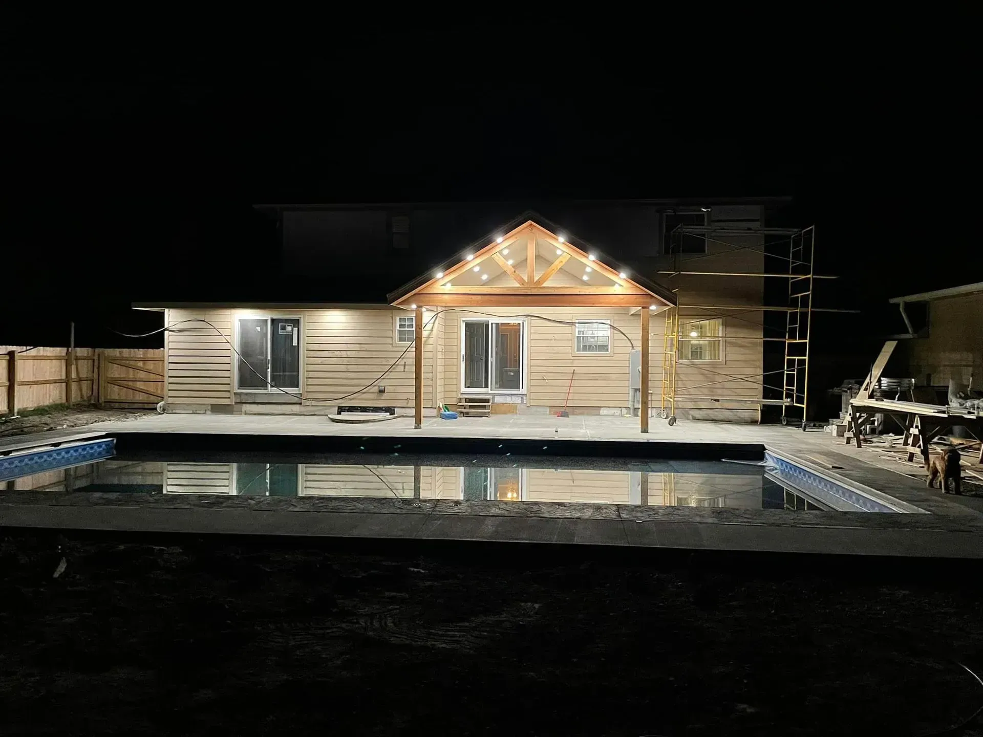 A house with a lit wooden porch and backyard pool at night, with construction scaffolding visible on the side.
