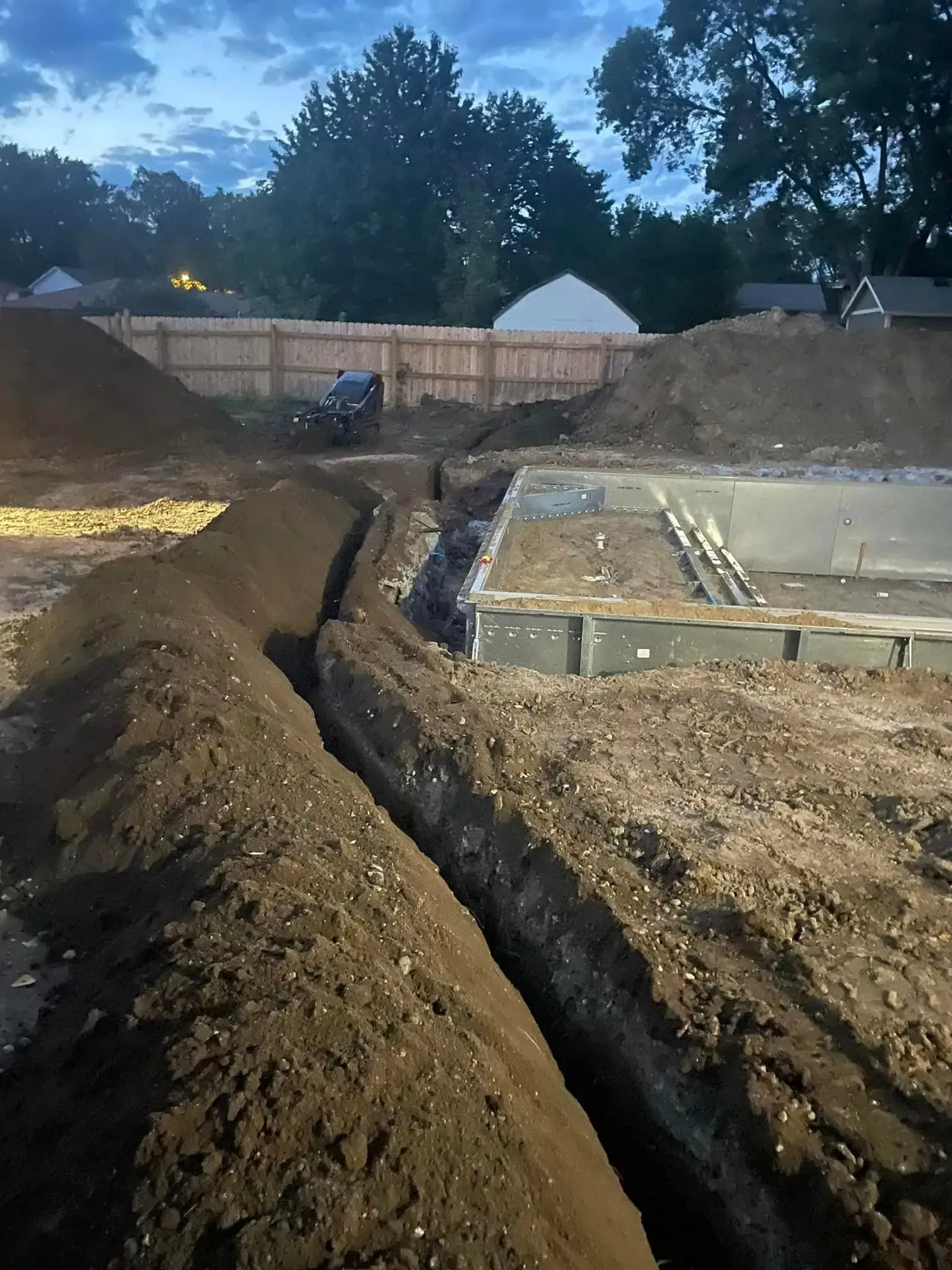 A deep, narrow trench runs through a construction site next to a concrete pool foundation under a dusky sky.