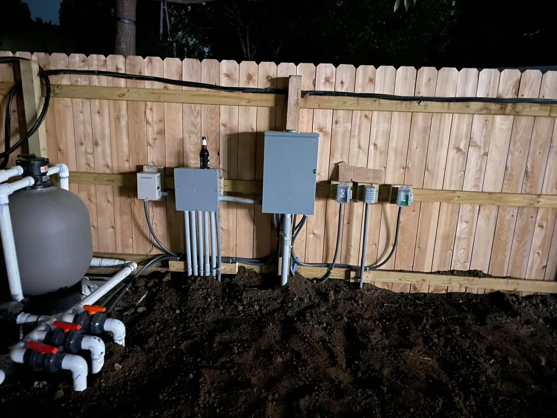 A pool filtration system with electrical panels and conduits mounted on a wooden fence outdoors.
