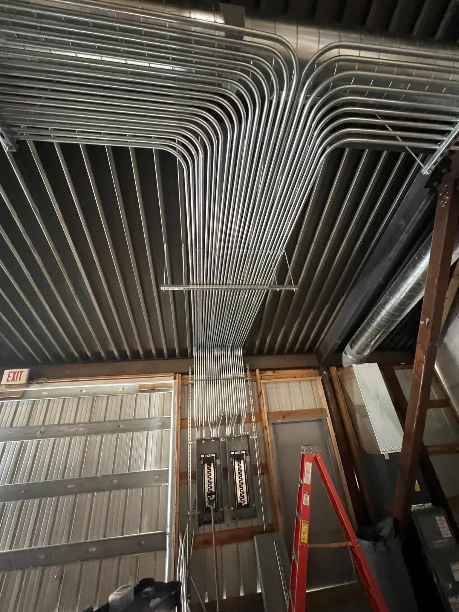 Rows of metal electrical conduits mounted to a corrugated ceiling run down into electrical panels on a wall.