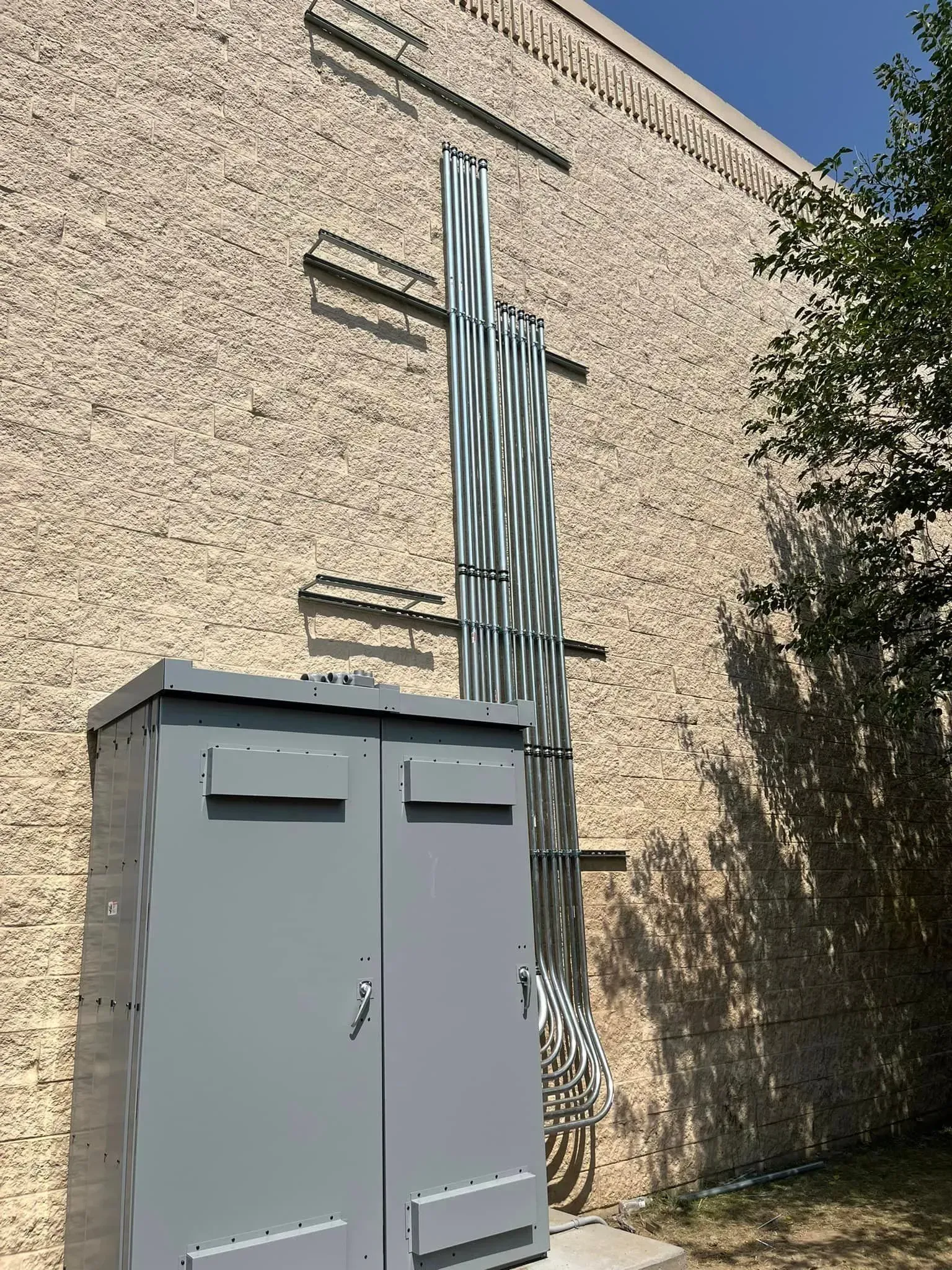 A grey utility cabinet mounted against a brick wall with several vertical metal conduits attached by horizontal supports.