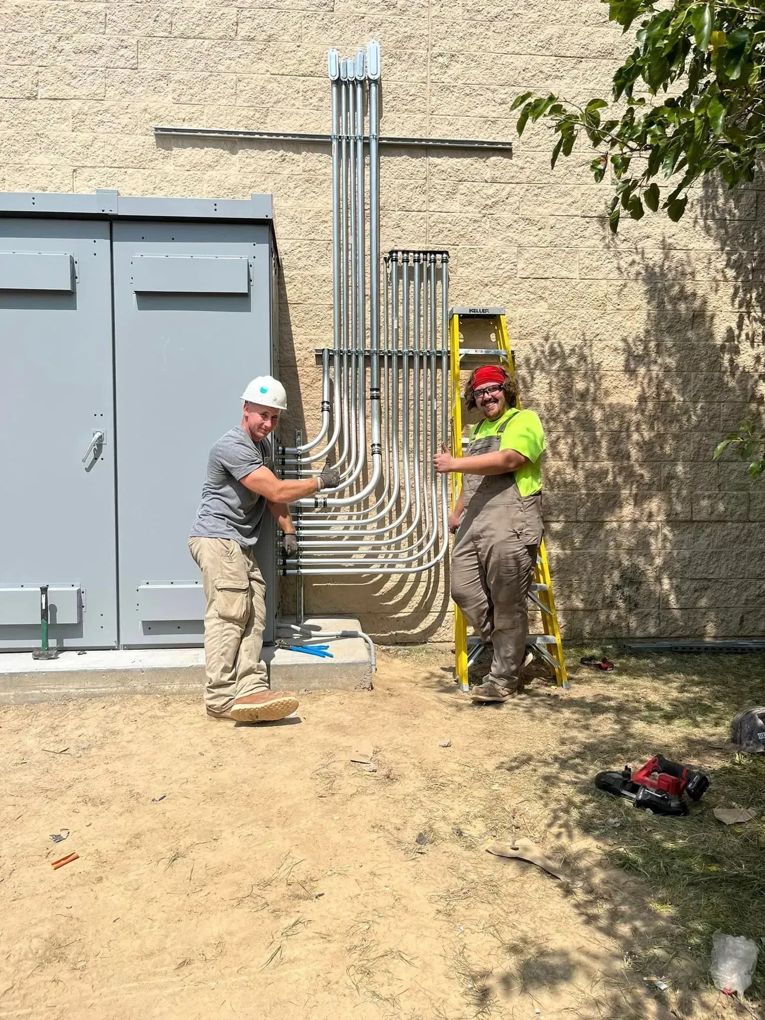 Two construction workers in high-visibility gear and hard hats pose next to a newly installed electrical conduit system.