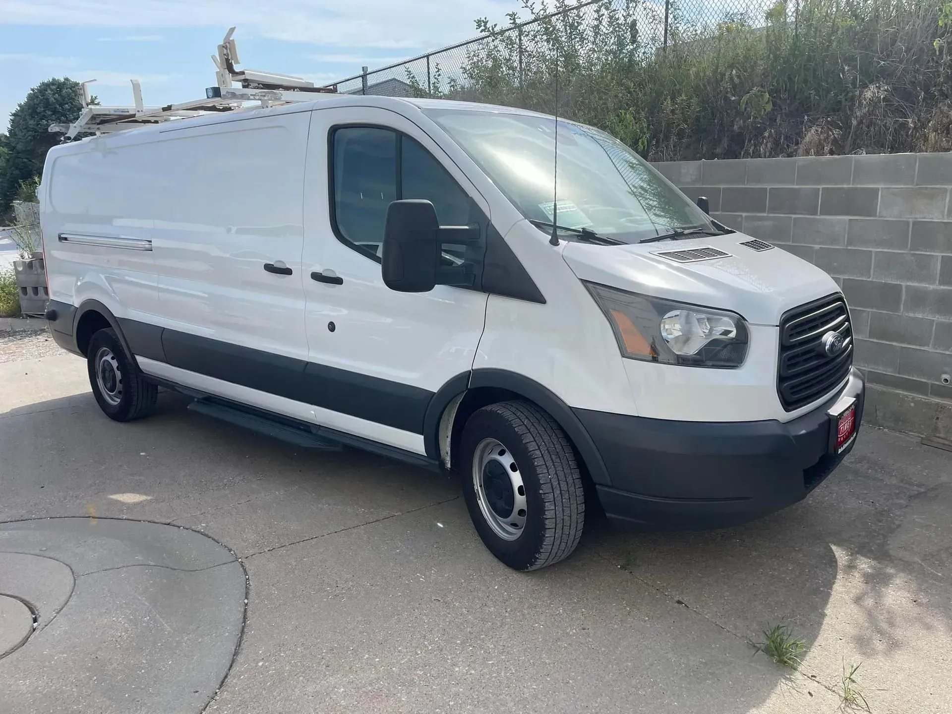 A white Ford Transit cargo van with a roof ladder rack parked on an asphalt lot near a concrete block wall.