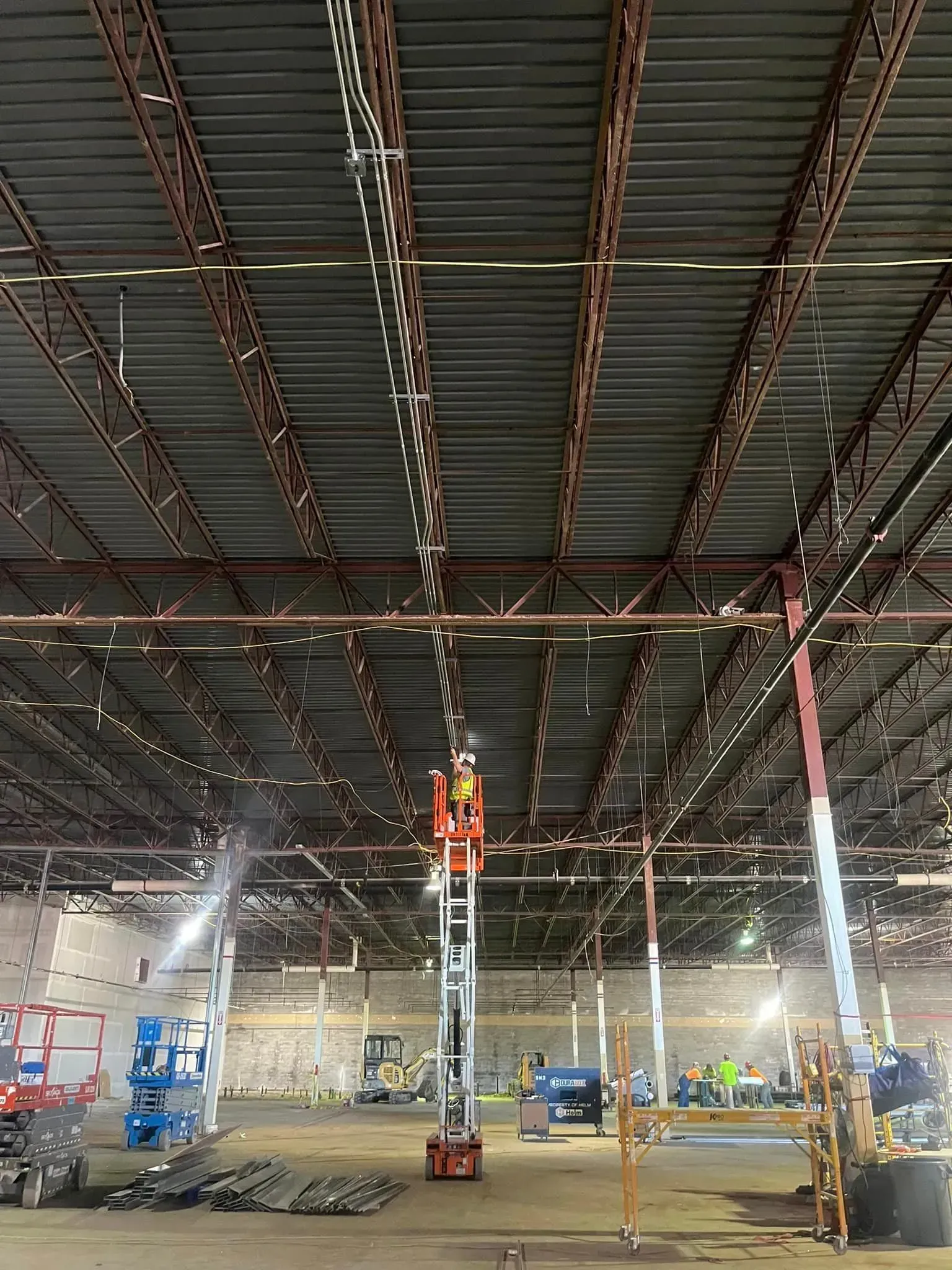 A construction worker on a vertical lift installs lighting in the high ceiling of a large, unfinished industrial space.