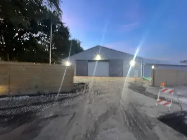 A warehouse building at dusk, featuring a large central door, illuminated by exterior lights, with a dirt foreground.