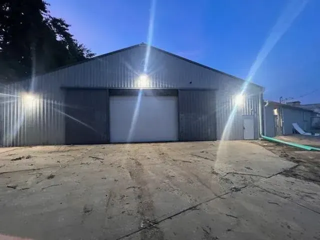A large, grey metal warehouse building with a closed central garage door and bright exterior lights at dusk.