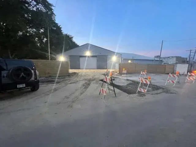 A construction site at dusk with a building, parked car, and orange-and-white safety barriers blocking a paved area.
