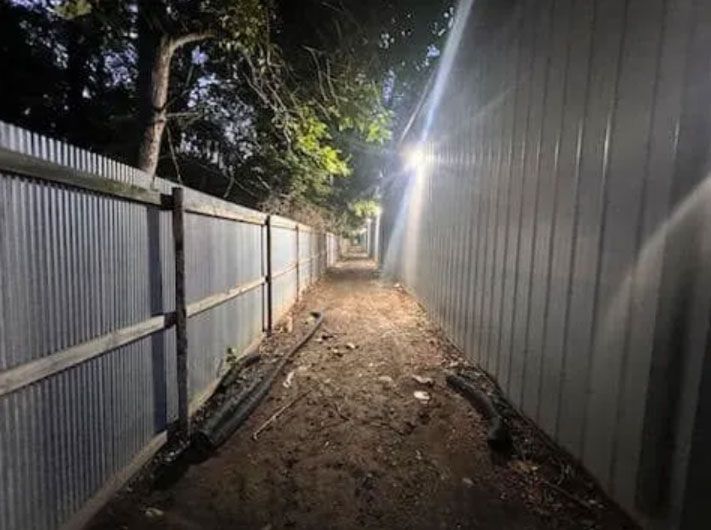 A narrow, dirt-floored alleyway at night, enclosed by high corrugated metal walls and illuminated by a bright overhead light.