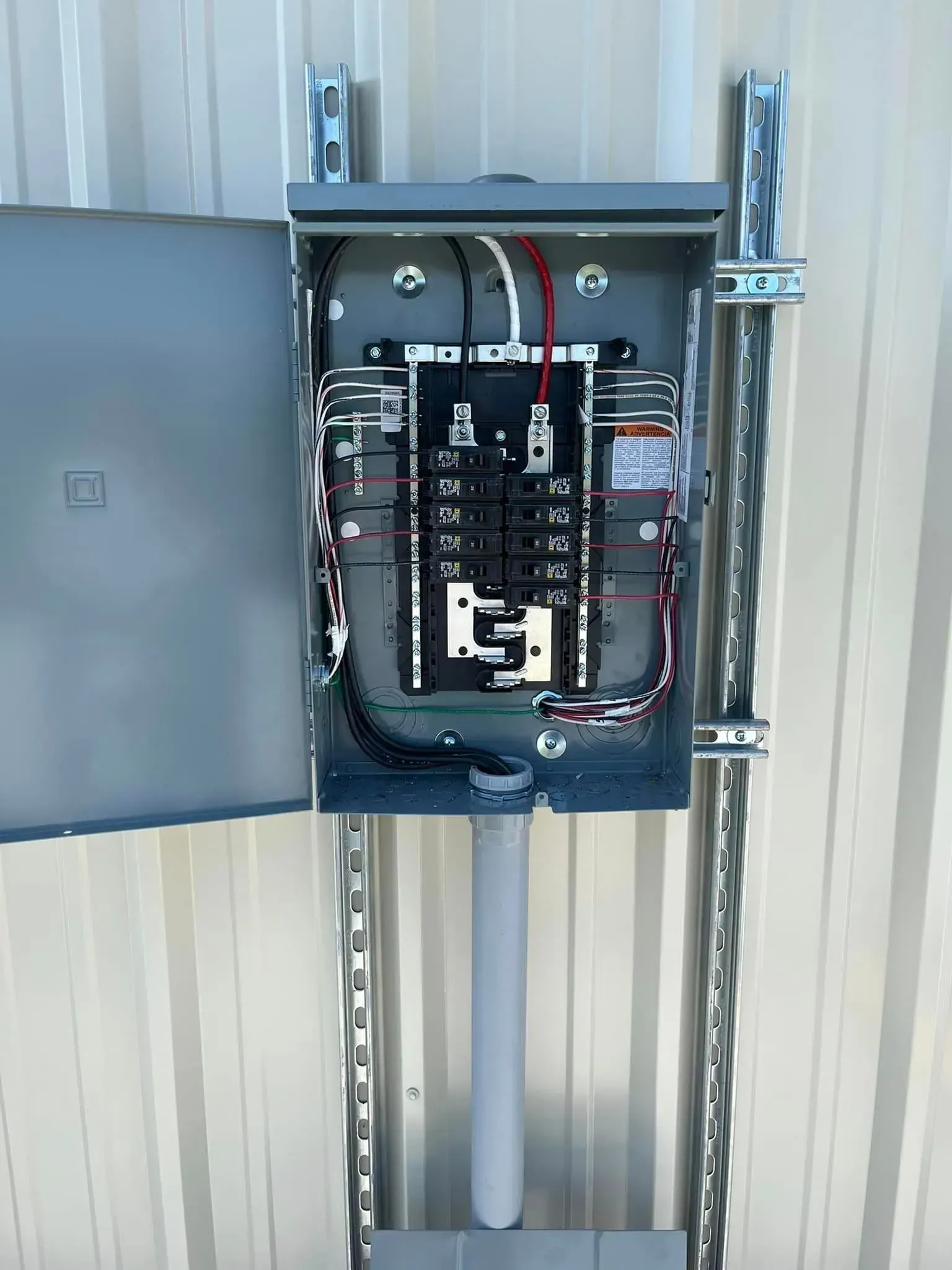 An open electrical breaker panel mounted on metal unistrut rails against a corrugated metal wall.