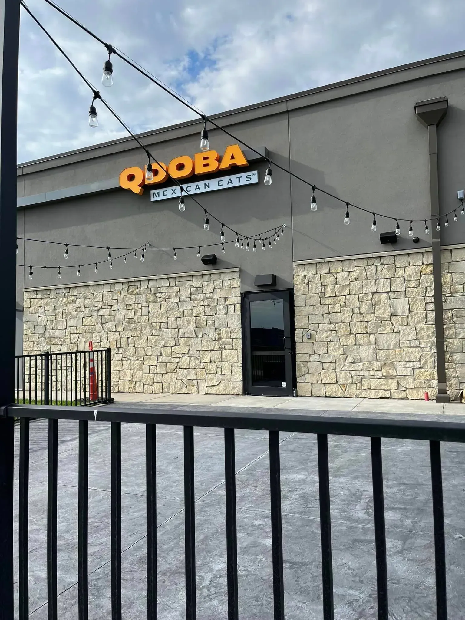 An exterior view of a Qdoba restaurant with a stone facade, a dark door, and string lights hanging across the front.