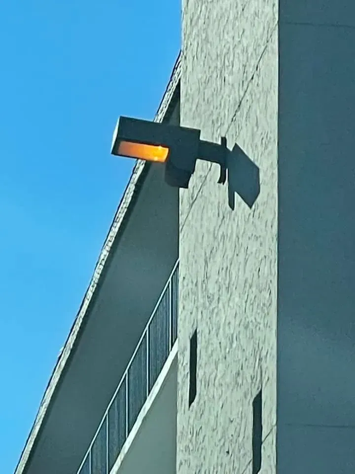 A square, illuminated exterior light fixture mounted on the side of a concrete building against a bright blue sky.