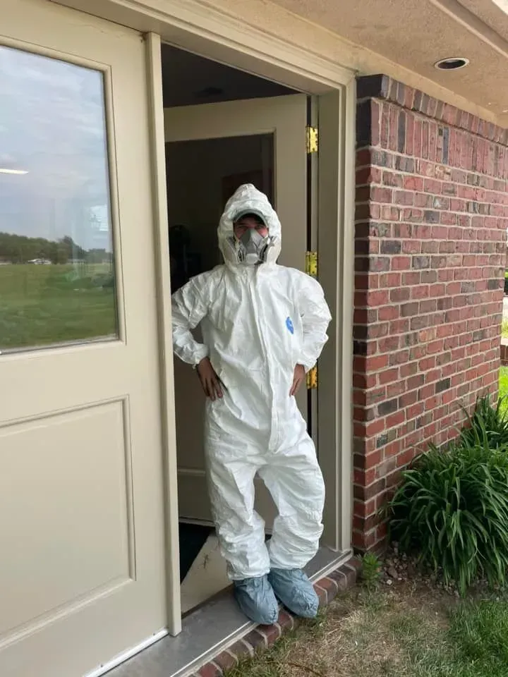A person in a white hooded protective suit and respirator mask standing in a doorway of a brick building.
