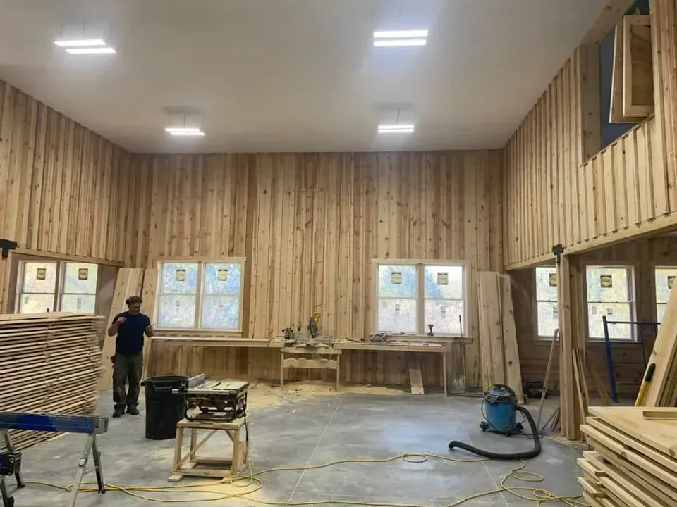 A person stands in a large room under construction with wood-paneled walls, concrete floors, and multiple windows.